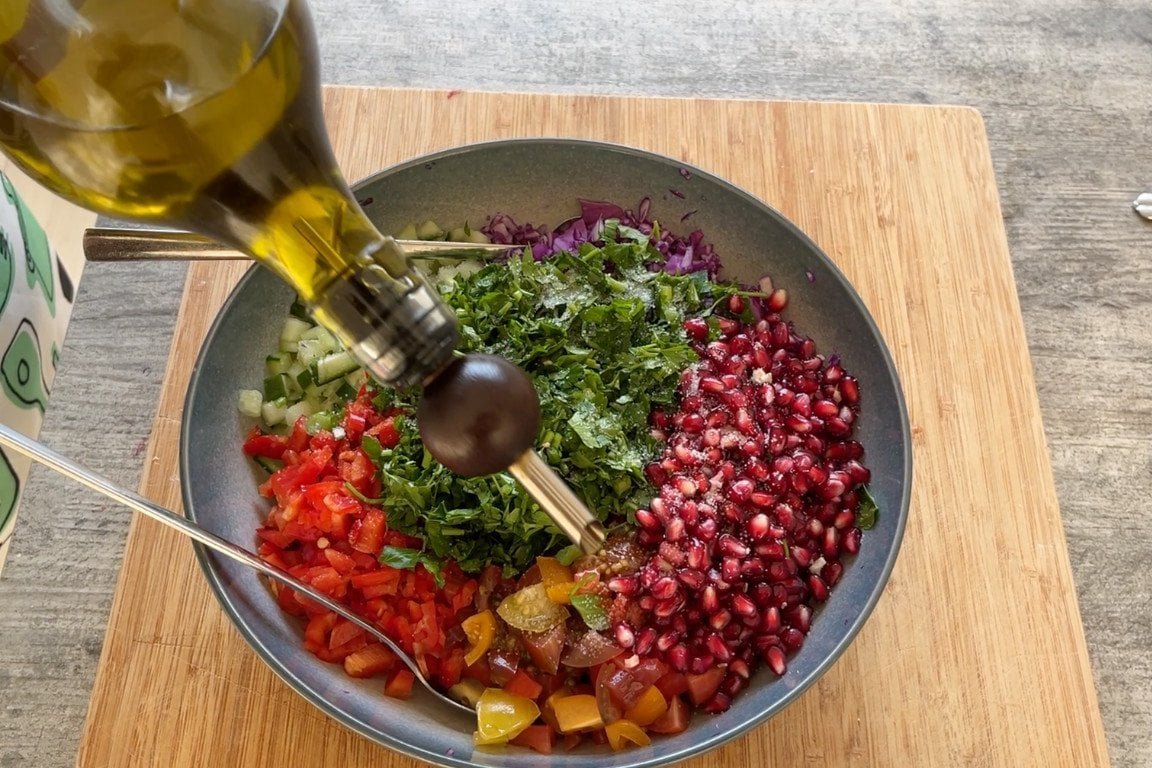 Een fles extra vierge olijfolie die over een rijkelijk gevulde salade met granaatappelpitjes, peterselie en gesneden groenten in een kom wordt gegoten