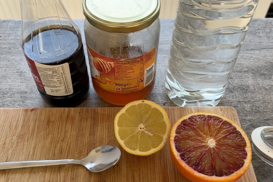 Verschillende ingrediënten voor een saladedressing op een houten ondergrond, waaronder flessen granaatappelmelasse, honing, azijn, gesneden citroen en een bloedsinaasappel