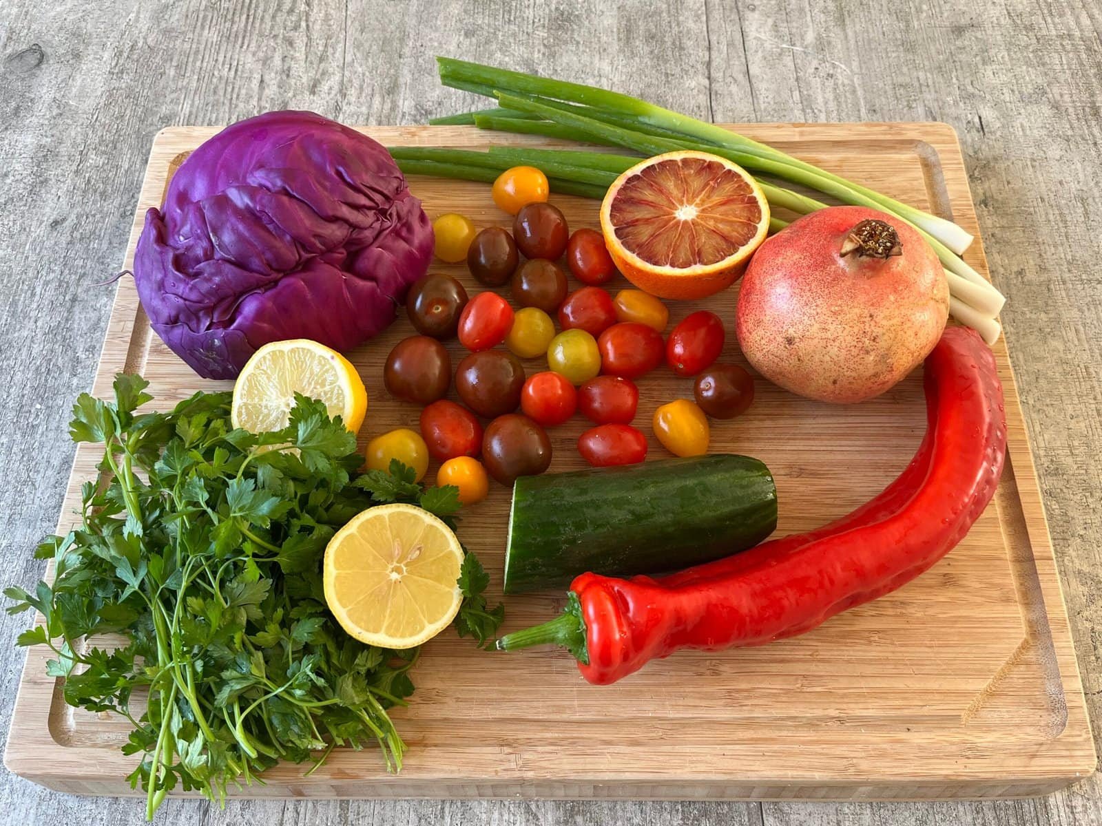 Diverse verse salade-ingrediënten op een houten snijplank, waaronder rode kool, cherrytomaten, granaatappel, peterselie en een rode puntpaprika
